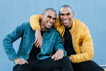 Portrait of African American twin brothers smiling and embracing each other, wearing blue and...
