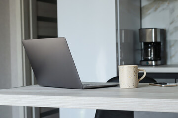 A laptop on a table with a cup, a phone on the table. on the background, a refrigerator, a coffee maker