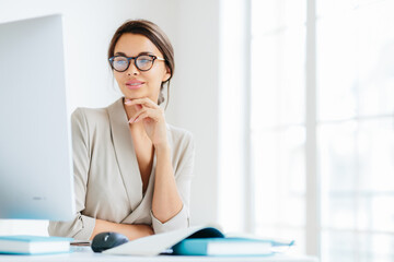 Charming female office worker keeps hand under chin, looks in screen of computer, wears spectacles for vision correction, poses in own cabinet, prepares information for future business meeting