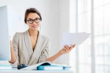 Indoor shot of good looking female in formal suit, holds paper documents, works on organization and creating startup project, poses at desktop with computer, checks information for presentation