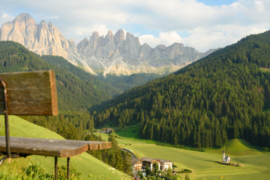 (Selective focus) Defocused park bench in the foreground with the Church of St. John (San Giovanni in Ranui) that stands out in the green meadows, in the heart of the beautiful Dolomitic landscape..
