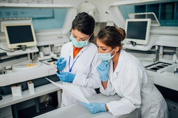 Two female scientists or technicians with face protective masks work in laboratory on human blood samples.