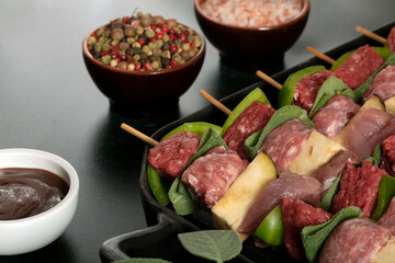 Macro close up of raw meat skewers before cooking on a dark background with sage leaves and mixed pepper.