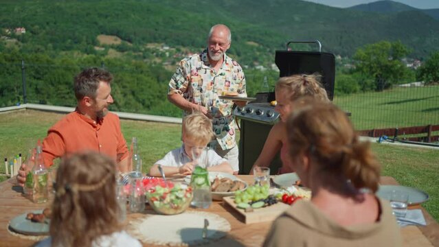 Happy senior man grilling and serving burgers at multi generation garden party in summer.