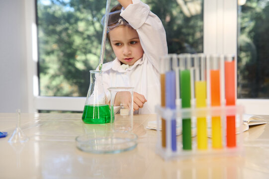 Focus on flat bottom flask with green color liquid chemical against background of pretty school girl using dropping bottle ground pipette, dripping reagent during experiments in chemistry class