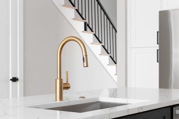 A kitchen sink detail shot with a gold faucet, grey island, white marble countertop, and lights...