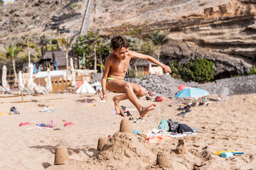 young man wearing a swimsuit jumping over a sand castle at an exotic beach near the sea