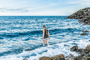 Fototapeta na wymiar Young woman with a pigtail in clothes enters the sea - rear view. Lonely girl against the backdrop of a seascape with big waves and a stone shore