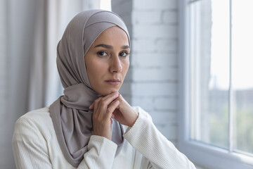 Portrait of young beautiful Muslim woman near window at home, Arab woman in hijab looking at camera with concentration, close up photo