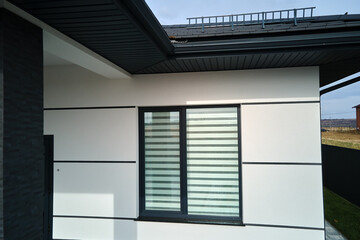 Corner of modern house with shingled roof and big window with closed shutters on white wall