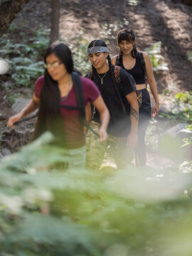 Vertical image of three Indigenous youth walking along a trail in a shaded forest 