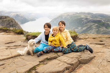 Fototapeta premium Family, enjoying the hike to Preikestolen, the Pulpit Rock in Lysebotn, Norway on a rainy day, toddler climbing with his pet dog the one of the most scenic fjords
