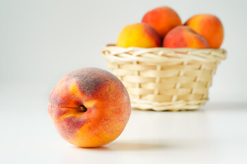 One ripe peach close-up in the foreground and several peaches in a basket behind it in blur on white background
