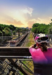 Una mujer vestida de ciclista y de espaldas observa el paso de un tren desde un puente.