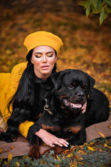 a brunette with a beautiful make-up in a yellow beret walks in autumn with a rottweiler and hugs a...