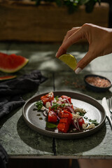 Squeezing lime juice above grilled watermelon salad with red onion and basil, dark mood photo. Focus on drops. Healthy food concept, copy space.
