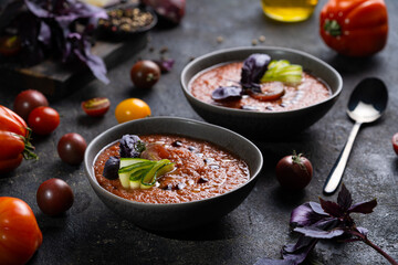Traditional spanish cold tomato soup gazpacho in a bowl served with basil and cucumber on black textured table with fresh ingredients around. Top view with copy space. Dark mood healthy vegan dish.