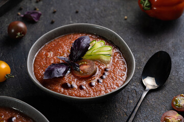 Traditional spanish cold tomato soup gazpacho in a bowl served with basil and cucumber on black textured table with fresh ingredients around. Top view with copy space. Dark mood healthy vegan dish.