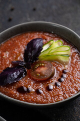 Traditional spanish cold tomato soup gazpacho in a bowl served with basil and cucumber on black textured table with fresh ingredients around. Top view with copy space. Dark mood healthy vegan dish.