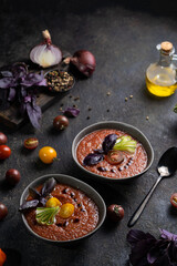 Traditional spanish cold tomato soup gazpacho in a bowl served with basil and cucumber on black textured table with fresh ingredients around. Top view with copy space. Dark mood healthy vegan dish.