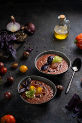 Traditional spanish cold tomato soup gazpacho in a bowl served with basil and cucumber on black textured table with fresh ingredients around. Top view with copy space. Dark mood healthy vegan dish.