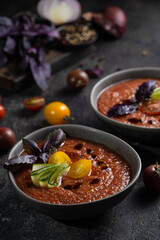 Traditional spanish cold tomato soup gazpacho in a bowl served with basil and cucumber on black textured table with fresh ingredients around. Top view with copy space. Dark mood healthy vegan dish.