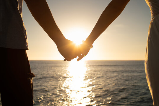 Couple bride and groom holding hands at sunset facing the ocean. Love and relationships concept. 