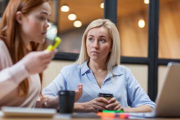 Two young women discussing business ideas while sitting at office desk with open laptop and working on project together. Focus on blonde woman with coffee listening to colleague explaining opinion