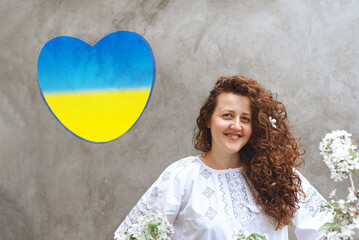 Woman with long curly hair in white embroidered dress. Blue and yellow heart on wall, like the State Flag of Ukraine. Defocused focus on foreground of flowers. Independence Day. Copy Space