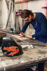 Side view of young metalworker in protective eyewear making marks on metal details with pencil before welding