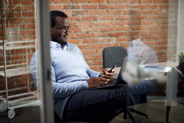 Black businessman using laptop while reading text message on cell phone in office.