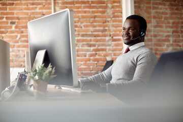 Happy African American call center agent wearing headset while working on computer in office.