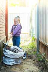 a little girl copies adults, she took a shovel and helps to clean up the garbage, old leaves in the yard of the house in a big trash bag