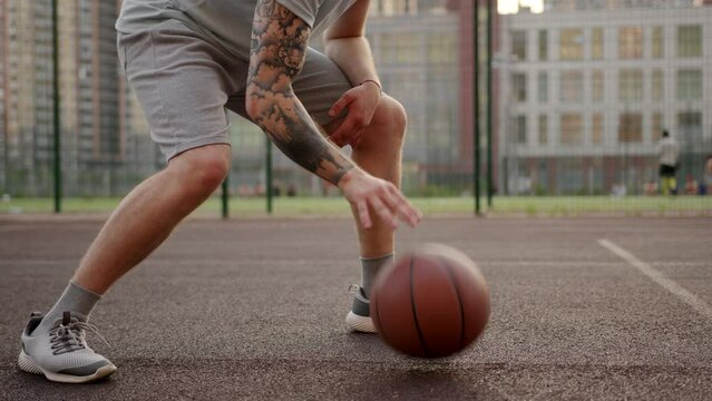 Sportsman in grey shorts bounces ball on basketball court against football field