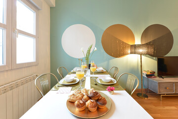 Dining room with rectangular white wooden table, metal chairs, muffins and pastries for breakfast and freshly squeezed orange juice