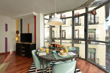 Dining room with a round wooden table with a large breakfast with fruits, juices and assorted pastries and a large glass and metal bay window with reddish wood floors
