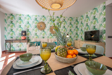 Dining room with wooden table with large breakfast area with fruits, juices and assorted pastries, living room with furniture and decoration on the walls