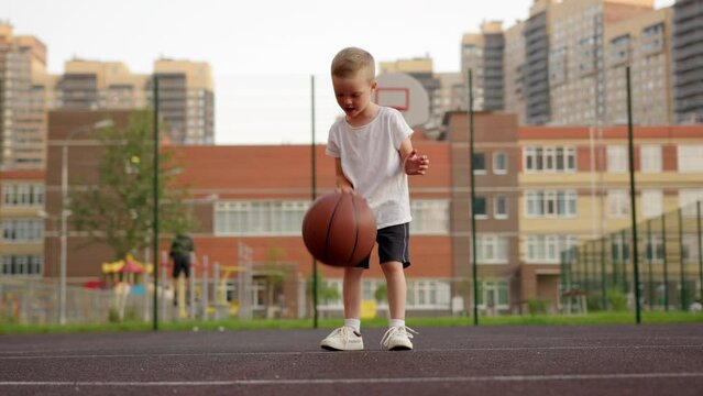Happy preschooler in white t-shirt and shorts bounces ball on basketball court