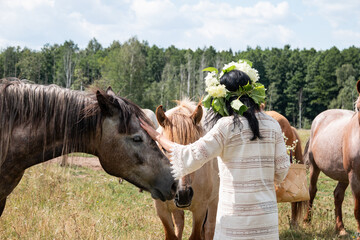 A girl in a wreath of flowers on her head and with a basket in her hands strokes a horse. A herd of...