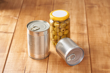 Iron tin can with tab opener and olives in a glass jar on the wooden table.