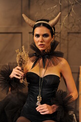 A beautiful brunette with horns and festive Halloween makeup with black manicure and black clothes sits with dry grass in her hands at home. Halloween makeup.