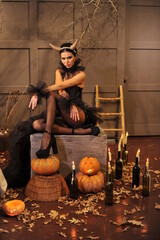 A beautiful brunette with horns and festive Halloween makeup with black manicure and black clothes sits on a chest in her house. Halloween makeup.