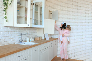 Mother prepearing breakfast together with dauther at home in kitchen. Smiling young caucasian woman in pajama sleep mask cooking food indoors with girl child on her arms. Toddler watching mobile phone