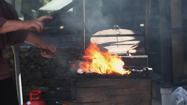 Middle-aged caucasian male blacksmith of the medieval era forges red-hot iron by pounding on it with a sledgehammer while standing in a forge on a summer day, close-up side view, slow motion.Artisanal