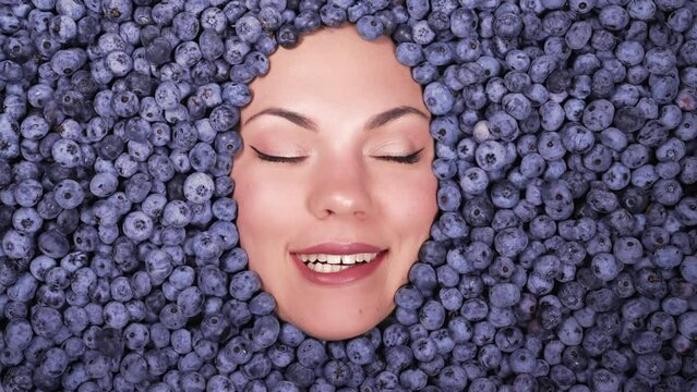 Woman licks lips, tempted by fresh ripe blueberries. Young attractive girl covered with acai berries. Lady with bright makeup winks hints at discounts. Advertising, diet, healthy vegan food. 