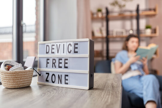 Digital Detox And Leisure Concept - Close Up Of Device Free Zone Words On Light Box, Gadgets In Basket On Table And Woman Reading Book At Home