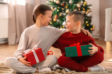 Obraz na płótnie Canvas christmas, winter holidays and childhood concept - happy girl and boy in pajamas with gifts sitting on floor and hugging at home