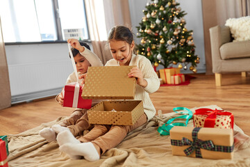 christmas, winter holidays and childhood concept - happy little girl and boy opening gifts sitting...