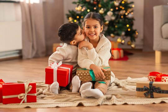 Christmas, Winter Holidays And Childhood Concept - Little Boy Kissing His Happy Sister Sitting On Floor With Gifts At Home