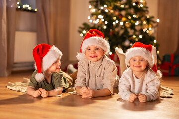 christmas, winter holidays and childhood concept - happy little girl and boys in santa hats lying on floor at home